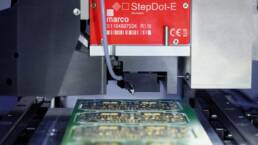 Close-up of the marco StepDot dispensing head above a green circuit board in a system for precise solder paste.